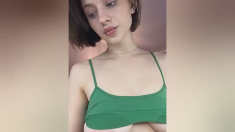 Media: Video of a young, fair-skinned woman with short, dark hair, wearing a green spaghetti strap crop top, standing against a blurred pink background.