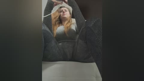 Media: Video of a blonde woman with fair skin, medium build, wearing a gray sweater and black knit leggings, lying on a white surface with her legs spread, looking distressed, possibly in restraints.