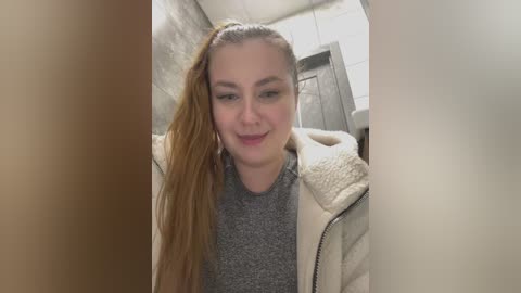 Media: A video of a smiling, fair-skinned woman with long, wet, blonde hair in a ponytail, wearing a grey sweater and beige jacket, standing in a bathroom with grey tiled walls and a white door.