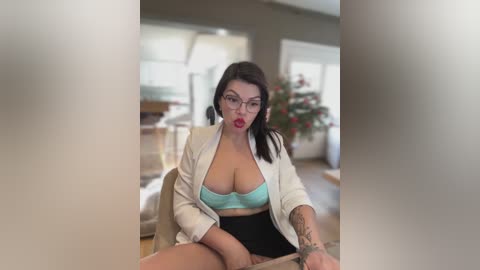 A video of a plus-size woman with glasses, fair skin, and dark hair, wearing a white blazer over a light blue bra, sitting in a modern living room.