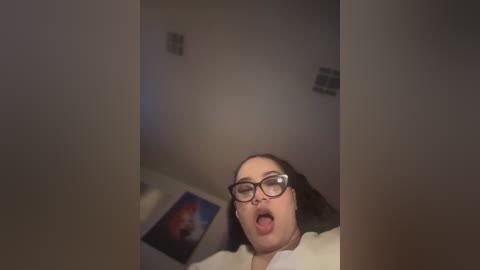 Media: Video of a chubby woman with glasses, tongue out, sitting in a dimly lit room with a colorful poster on the wall.