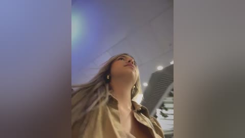 Video of a young woman with long, blonde hair, wearing a beige shirt, gazing upwards. Background features blurred, modern indoor setting with light blue and white tones.