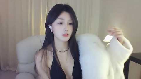 Media: A video of a young East Asian woman with long black hair, fair skin, and small breasts, wearing a black halter top, sitting on a white chair, looking at her phone.