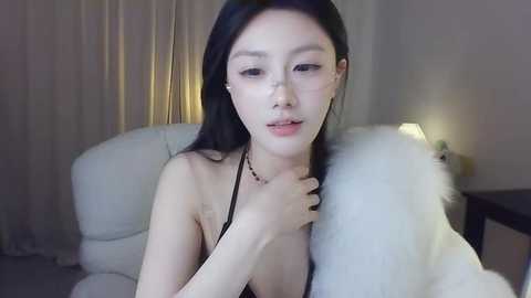 Media: Video of an East Asian woman with long black hair, fair skin, and small breasts, wearing a black strapless top, seated on a white fur chair in a dimly lit room.