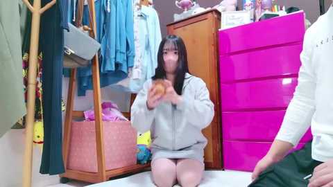 Media: Video of a young Asian woman with long black hair, wearing a grey hoodie and pink skirt, kneeling in a cluttered bedroom with vibrant colors and stuffed toys.