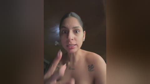 Media: Video of a young Latina woman with medium brown skin, dark hair pulled back, topless, holding a phone, in a dimly lit room with wooden walls.