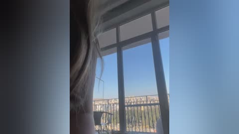 Video of a person's long hair blowing in the wind near a balcony railing. The background shows a clear blue sky and a cityscape with buildings and greenery.