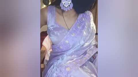 Media: Video of a woman in a light purple, intricately embroidered saree with a white floral headpiece, seated indoors with blurred background.