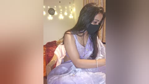 Media: Video of a young South Asian woman in a light purple saree, black mask, and long black hair, standing indoors near a bed with red and yellow bedding.