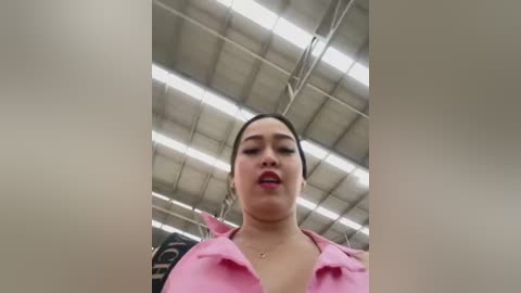 Media: A low-angle video of a Latina woman with medium skin tone, dark hair, and pink lipstick, wearing a pink shirt, standing in a warehouse with fluorescent lighting and exposed beams overhead.