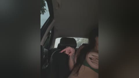 Media: Video of a woman in a car, wearing green lingerie, covering her face with her hand, sitting in a dark interior, with a blurry window view of trees.