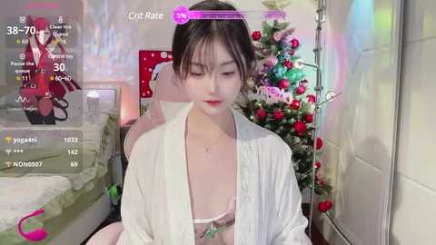 Media: Video of an Asian woman with fair skin, dark hair, and a slender figure, wearing a white robe, sitting in a cozy room with a decorated Christmas tree and gaming elements.