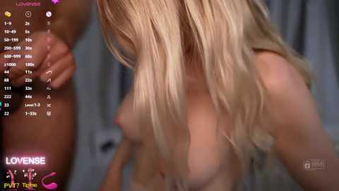 A close-up video of a nude, blonde woman with fair skin and medium-sized breasts, taken in a dimly lit room with blurred background. The image includes a digital timer display in the upper left corner.