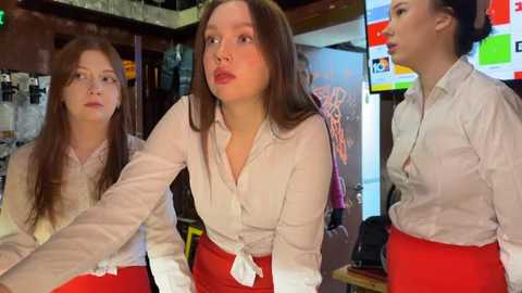 Media: Video of three young women in a cozy, dimly-lit room, wearing white blouses and red skirts, looking at a TV screen displaying colorful images.