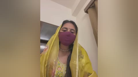 Media: A video of a woman in a yellow sari with a purple mask, standing in a dimly lit room. The image is slightly blurred.