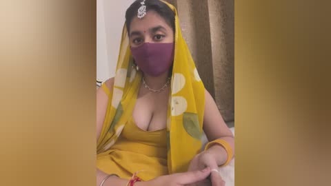 Media: Video of a woman with medium skin tone, wearing a yellow sari with white polka dots, a purple mask, and a silver nose ring, seated against a beige curtain backdrop.