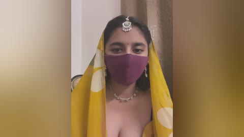 Media: A video of a young South Asian woman with a light brown complexion wearing a purple face mask, yellow polka-dotted saree, and a traditional headpiece. She is indoors with beige curtains in the background.