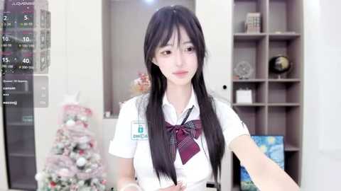 Media: A video of a young Asian woman with long black hair, wearing a white school uniform with a red plaid bow tie, standing in a modern, brightly lit room with a Christmas tree and bookshelves in the background.