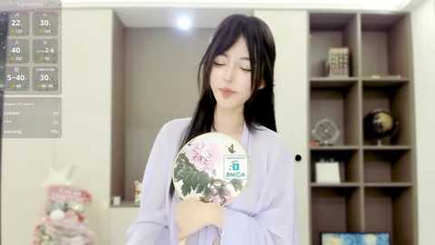 Media: A video of an East Asian woman with long black hair, wearing a light lavender blouse, holding a pink flower bouquet. She stands in a modern, beige-toned room with a digital clock on the left and a bookshelf on the right.