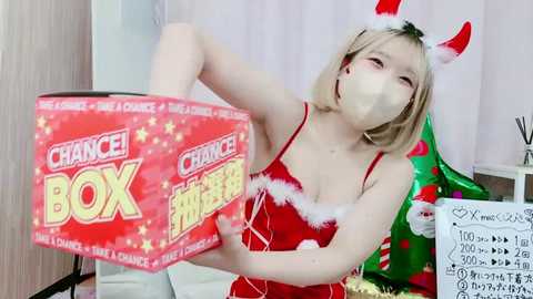 Media: Video of a young, fair-skinned Asian woman with blonde hair, wearing a red Santa dress and a Christmas-themed mask, opening a red \"Chance! Christmas Box\" box, in a festive room with holiday decorations.