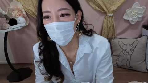 Media: Video of an Asian woman with long, wavy black hair, wearing a white surgical mask, white button-up shirt, and gold earrings. Background features pink and beige decor, including a table with stuffed toys and a beige pillow with a pug design.