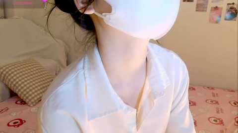 Media: Video of a person with light skin, wearing a white mask, white shirt, and a patterned pink and white bedspread in a softly lit bedroom.