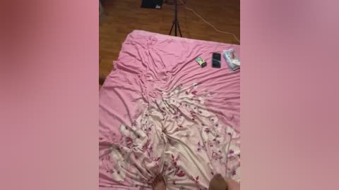 Media: Video of a messy bedroom with a rumpled pink bedspread, a smartphone, and a power cable on a wooden floor, suggesting a disorganized living space.