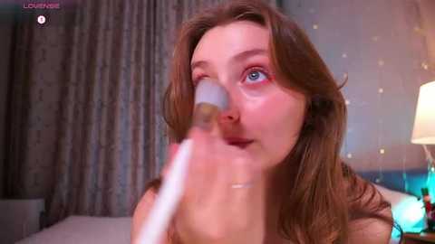 Media: A video of a fair-skinned woman with brown hair, wearing a white top, applying makeup to her face with a brush in a softly lit bedroom with a bed, lamp, and curtains.