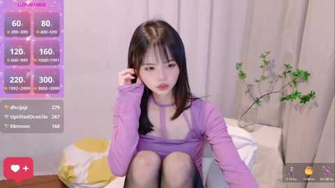 Media: A video of a young East Asian woman with fair skin and straight black hair, wearing a purple top, sitting on a bed with a plant in the background.