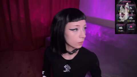 Video of a pale-skinned, black-haired young woman with a gothic look, wearing a black choker and long-sleeved top, in a dimly lit room with purple and red lighting.