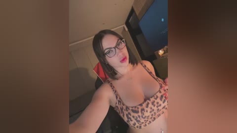 Media: Video of a fair-skinned woman with short black hair and glasses, wearing leopard-print lingerie, sitting in a dimly lit room with a window.