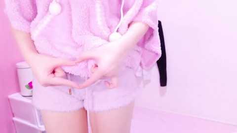 Media: A video of a young, fair-skinned individual in pastel-colored, fuzzy pink pajamas, wearing white lace panties, adjusting them. The background is a soft pink wall and white floor, with a white shelf on the left holding a white can.