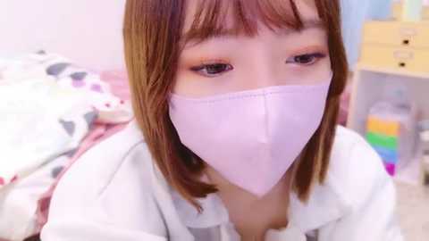 Media: A video of an East Asian woman with light skin, shoulder-length brown hair, and a white face mask, wearing a white shirt, sitting on a bed with a colorful, patterned blanket. The background includes a dresser with colorful items and a white wall.