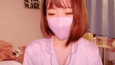 Media: Video of an Asian woman with short, straight brown hair, wearing a lavender mask, a pink robe, and a pink pillowcase in a bedroom with pastel colors and a \"OF THE PLACE\" sign.