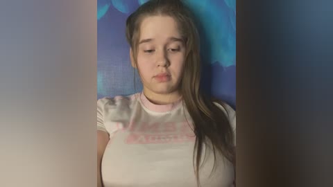 Media: Video of a young woman with light skin, brown hair, and a slightly puffy face, wearing a light pink t-shirt with \"MILK\" in white text. She stands against a blue and purple abstract background.