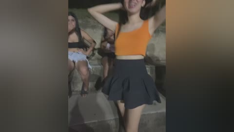 Media: A blurry, low-quality video of three young women in casual attire, one in a black top and shorts, another in an orange top and black skirt, and the third in a white top and shorts, sitting on a concrete surface.