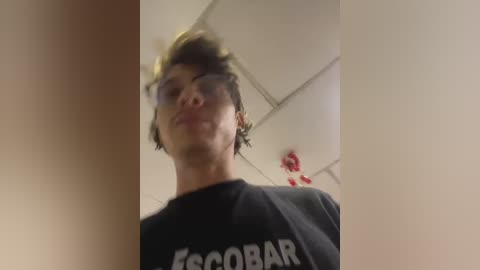 Media: Video of a young man with short, messy hair, wearing glasses and a black \"Escobar\" t-shirt, captured from a low angle in an indoor setting with fluorescent lights.