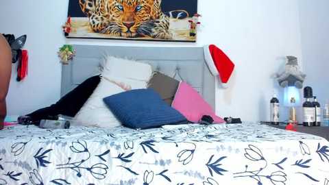 Media: Video of a modern bedroom featuring a bed with a white floral-patterned duvet, pillows in various colors, a framed tiger print above, and festive decorations including a Santa hat and red and white balloons.