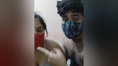 Media: A video of a man with dark curly hair and a woman with long dark hair, both wearing face masks, holding a red phone in a dimly lit room.