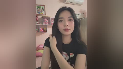 Media: Video of an Asian woman with long black hair, wearing a black T-shirt, in a pink room with stuffed animals, shelves, and air conditioner.