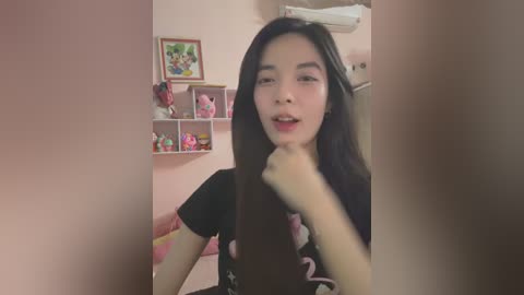 Media: Video of an Asian woman with long black hair, wearing a black top, making a \"victory\" gesture in a pink bedroom with shelves and plush toys.