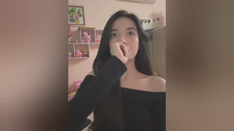 Media: Video of an Asian woman with long black hair, wearing a black off-shoulder top, covering her mouth with her hand, in a pink-walled room with shelves and toys.