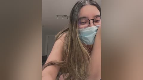 Media: Video of a young woman with long, straight brown hair, wearing a blue surgical mask and black-framed glasses, leaning against a beige wall.