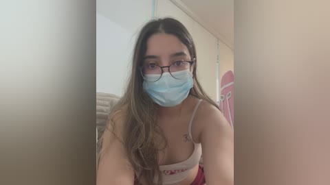 Media: A video of a young woman with long, wavy brown hair, wearing a blue mask, black-rimmed glasses, and a white tank top, sitting in a beige room with a pink poster.