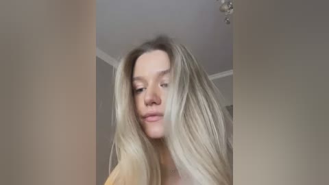 Media: A video of a young Caucasian woman with long, blonde hair, light skin, and a slim figure, wearing a beige top, standing indoors in a softly lit room with a chandelier visible.