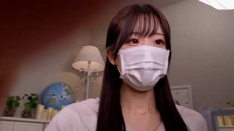 Media: Video of an East Asian woman with long black hair, wearing a white surgical mask, standing in a modern, brightly lit room with a lamp, globe, and white cabinets in the background.