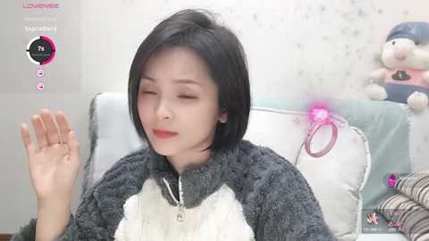 Media: Video of an East Asian woman with straight black hair, wearing a gray panda-themed hoodie, sitting on a white bed, with a pink heart floating beside her head.