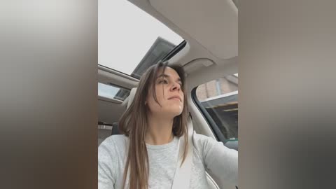 Media: Video of a young woman with long brown hair, light skin, and a relaxed expression, seated in a car with a beige interior.