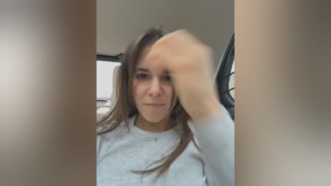 Media: Video of a young woman with straight, shoulder-length brown hair, wearing a light gray long-sleeved top, sitting in a car with the door open, partially obscured by the frame, giving a subtle, introspective expression.