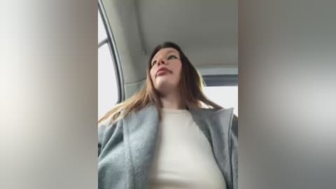 Media: A video captures a woman with long, straight brown hair, wearing a gray blazer over a white shirt, looking up through a car window. The background is a dimly lit, minimalist interior.